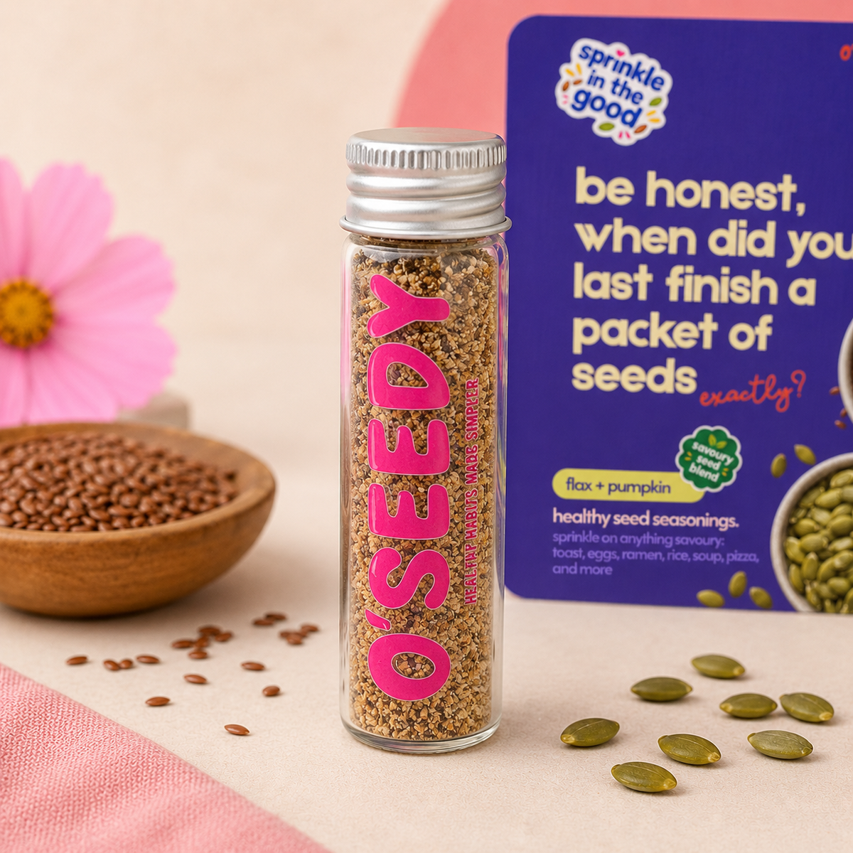 A clear glass bottle of OSEEDY pumpkin and flax seed seasoning stands in front of a purple branded package. The bottle contains tan-brown seeds and has bold pink lettering.