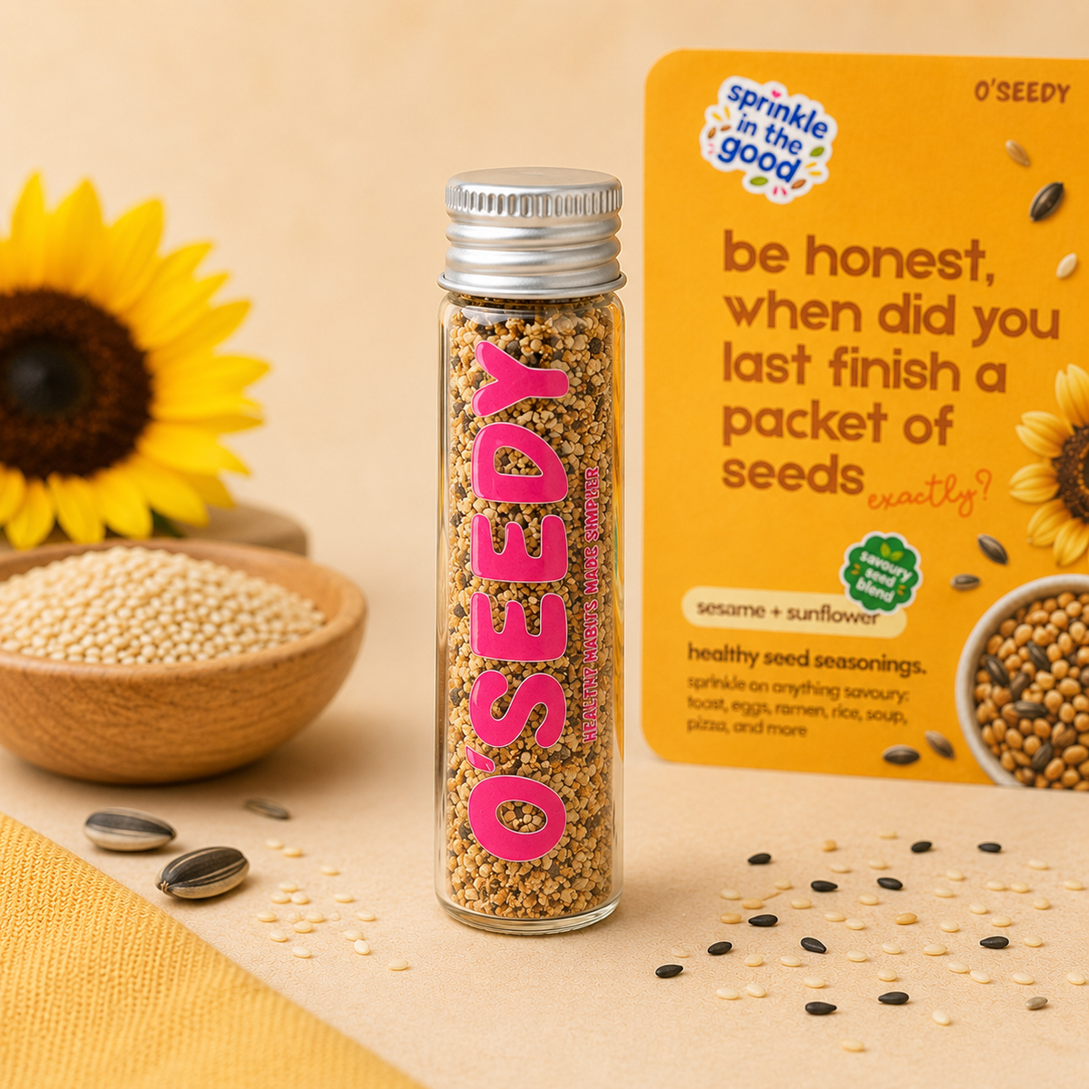 A glass bottle of O'Seedy Sprinkle in the Good Sesame + Sunflower seasoning stands on a beige surface next to its yellow packaging card.